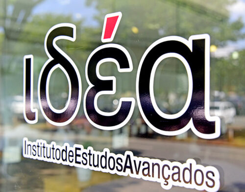 Na imagem, a escrita IdEA, em preto com contorno branco, colada no vidro que reflete uma paisagem com folhas verdes ao fundo.
