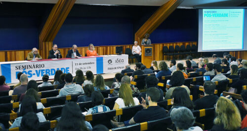 Audiodescrição: No palco de um amplo auditório, imagem em perspectiva e panorâmica, quatro pessoas sentadas em cadeiras, uma ao lado da outra, sendo que a segunda à esquerda fala em microfone sem fio. Há uma extensa mesa retangular à frente deles, com um banner afixado à frente, onde se lê Seminário Pós-Verdade. À frente deles, parte do público do auditório, cerca de sessenta pessoas que se encontram sentadas em cadeiras dispostas uma ao lado da outra, formando várias fileiras horizontais. Imagem 1 de 1.